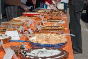 A long table covered with an orange cloth displays many different pies, some labeled with cards. During September in Bigfork, several people, mostly out of frame, are serving pieces of pie using utensils.