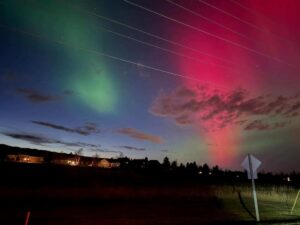 Colorful northern lights in green and red illuminate a winter night sky above houses and trees, with power lines stretching across the scene and a road sign visible in the foreground.