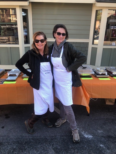 Two women wearing white aprons and sunglasses stand smiling with their arms around each other in front of a table covered with an orange tablecloth and food trays, celebrating an Autumn outdoor gathering in front of a building.