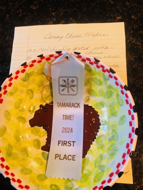 A yellow plate with a brown bear design holds a white ribbon that reads, TAMARACK TIME! 2024 FIRST PLACE. Behind the plate is a handwritten recipe titled Cheesy Cheese Wafers—an autumn celebration worth savoring.