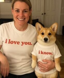 A smiling woman and her corgi sit side by side in matching white shirts at Bigfork Days. The woman's shirt says i love you, while the dog's reads i know. in red letters.
