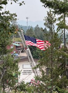 An American flag hangs from a fire truck ladder above the Bigfork Fire Dept. building, surrounded by trees as fall brings cool crisp days, with mountains and a cloudy sky in the background.