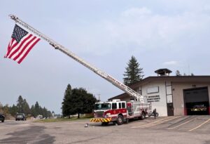 A fire truck with an extended ladder displays a large American flag in front of a fire station. The truck is parked on a paved lot, and the scene features trees showing signs of changing season, a cloudy sky, and a few parked vehicles.