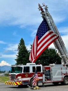 A large American flag hangs from a fire truck’s extended ladder in Bigfork. The truck is parked outdoors among trees and mountains, with firefighter gear and a small American flag placed in front—a proud September scene in the fall.