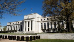A large, white neoclassical building with columns at the entrance, a U.S. flag flying on the roof, surrounded by trees and blooming homes under a clear blue sky.