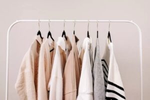 A white clothing rack displays seven neutral-toned garments perfect for the spring season, including beige, cream, white, and gray tops, all hanging neatly on black and gold hangers against a light wall.
