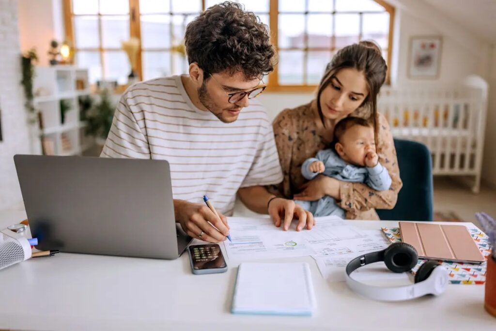 A man and woman sit at a desk with financial papers, a laptop, and a calculator. Embracing Spring Success, the woman holds a baby while the man writes. The focused family works in a bright home office with a crib in the background.