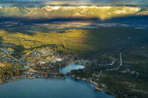 Aerial view of a forested area with a small town by a lake, surrounded by pine trees and mountains with patches of snow, under a partly cloudy sky with sunlight casting shadows.