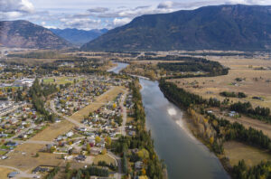 A river winds through a rural town with houses on one side and open fields on the other, bordered by forested hills and distant mountains under a partly cloudy sky.