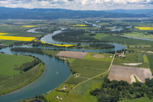 A winding river flows through a patchwork of green and yellow farmland, with distant mountains and clouds in the background under a bright sky.
