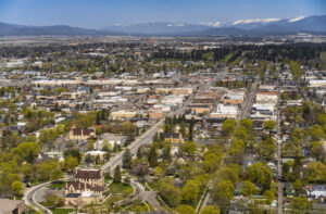 Aerial view of a small city with a grid layout, tree-lined streets, historic buildings, and mountains with snow-covered peaks in the background under a clear blue sky.