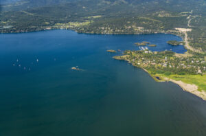 Aerial view of a large blue lake surrounded by green forested hills, with small islands, sailboats on the water, and a shoreline with scattered buildings and grassy areas.