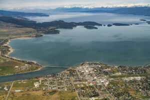 Aerial view of a small town by a large lake with a bridge, surrounded by green fields, forests, and distant snow-capped mountains under a partly cloudy sky.