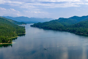 A wide, calm river winds between densely forested hills and mountains under a partly cloudy sky, with reflections of trees and clouds visible on the waters surface.