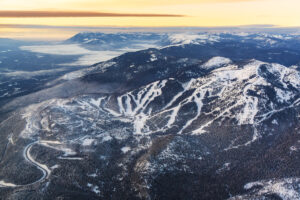 Aerial view of a snow-covered mountain ski resort with winding ski runs and forested slopes under a golden sky at sunrise or sunset, surrounded by distant mountains.